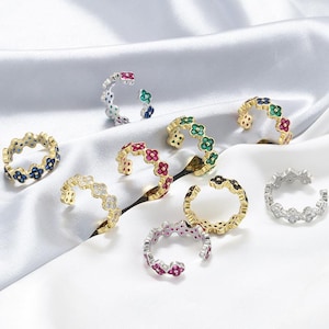 May include: A collection of open-band rings featuring a repeating four-petal flower motif. Each ring is crafted with metallic bands and inlaid with small, colorful gemstones including blue, red, green, and clear stones in various metal finishes.