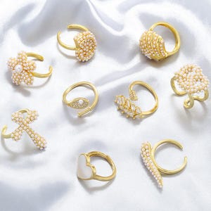 May include: A collection of gold-toned rings with various designs, including floral, heart, and abstract shapes. Many rings are adorned with small, white pearls, creating a delicate and elegant aesthetic. The rings are displayed on a white, silky fabric.