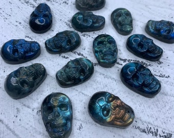 Puede incluir: Una colección de 14 cabujones en forma de calavera de color azul y verde iridiscente. Los cabujones están hechos de labradorita y tienen un efecto brillante único.