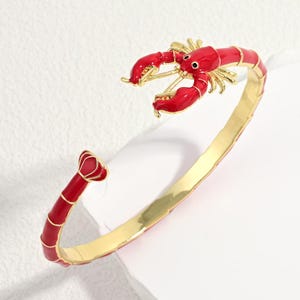 May include: A gold-tone open cuff bracelet featuring a red enamel lobster motif. The lobster design forms the focal point of the piece, with the tail section at one end and the claws at the other, connected by a smooth, polished gold-colored band.