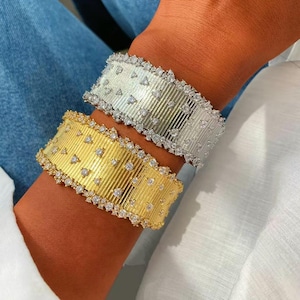 May include: Two wide cuff bracelets, one in silver tone and one in gold tone, featuring a vertical ribbed texture across the surface. Both bracelets are embellished with scattered, small, clear, round, sparkling stones along the ribbed bands.