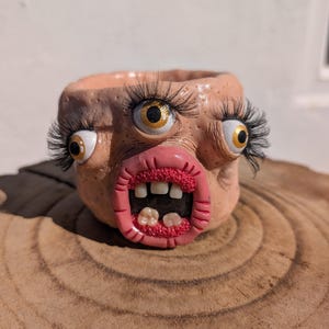 Könnte beinhalten: Ein pfirsichfarbener Keramik-Pflanztopf in Form eines Gesichts mit drei gelben Augen, langen schwarzen Wimpern und einem weit geöffneten Mund mit rosa Lippen und mehreren weißen Zähnen. Die Oberfläche hat eine texturierte, hautähnliche Optik.