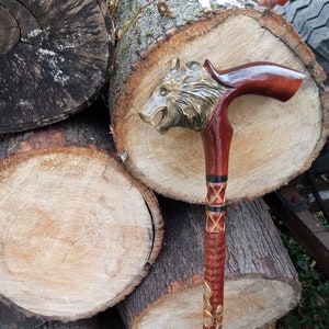 Op de afbeelding: Een houten wandelstok met een gegraveerd wolfskop handvat. Het handvat is bruin en goud geverfd, en de schacht is versierd met gegraveerde bladeren en een zwart-wit patroon.