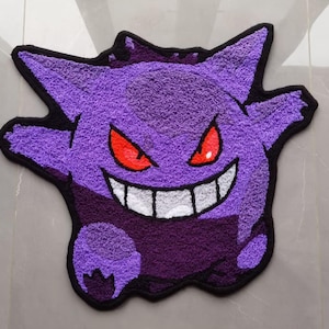 May include: A tufted rug shaped like the character Gengar, featuring a vibrant purple body with a darker purple lower section. The design includes sharp, pointed ears, glowing red eyes, and a wide, toothy grin outlined in black yarn.