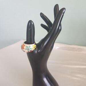 May include: A white ring with a colourful floral design. The ring is made of metal and has a smooth, rounded surface. The flowers are painted in shades of orange, yellow, green, and blue.