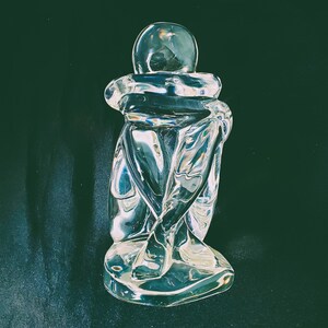 May include: A clear glass sculpture of a person in a seated position, hugging their knees. The figure has a rounded head and is made of transparent glass, reflecting light. The sculpture sits on a small base.