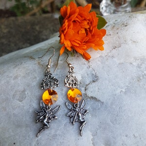 Op de afbeelding: Een paar zilveren oorbellen met oranje kristallen druppels en een fee bedel aan elke oorbel. De oorbellen hebben een filigraan ontwerp.