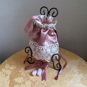 May include: A dusty rose satin drawstring pouch featuring a white floral lace overlay, a matching satin ribbon closure, and two small fabric flower accents at the base. The pouch is displayed on a dark brown metal scrollwork stand.