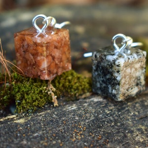 May include: Two small, cube-shaped pendants with silver loops. The pendant on the left is filled with brown and orange flecks, while the pendant on the right is filled with black and white flecks.