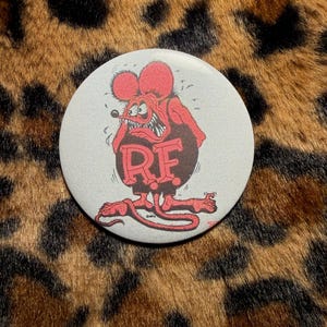 May include: A round button featuring a cartoon rat character in red and black. The rat has an angry expression and the letters "RF" are prominently displayed on its torso. The background is a light gray color.