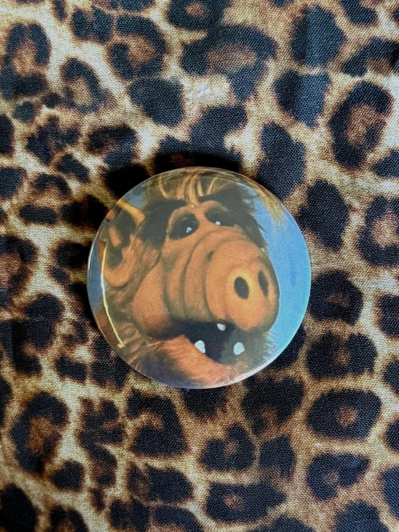 May include: A circular pinback button featuring a close-up portrait of the character Alf, a brown, furry extraterrestrial with a large snout, dark eyes, and a wide, open mouth showing two white teeth against a light blue background.