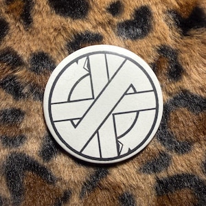 May include: A round, white button with a black, geometric design. The design features intersecting lines and shapes, set against a leopard print background. The button is a simple, graphic accessory.