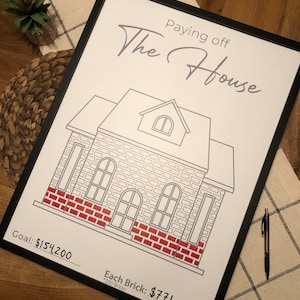 May include: A framed print with the text "Paying off The House" above a house illustration. The house has red brick accents. The goal is $154,200, with each brick costing $771. A black pen rests on the table.