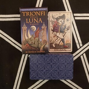 May include: A tarot card deck with a blue box and a card featuring an illustration of a figure labeled "THE FOOL." The box reads "TRIONFI DELLA LUNA TAROT" and "BY PATRICK VALENZA."