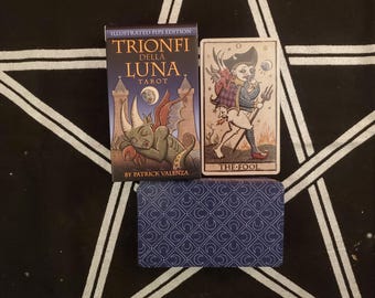 May include: A tarot card deck with a blue box and a card featuring an illustration of a figure labeled "THE FOOL." The box reads "TRIONFI DELLA LUNA TAROT" and "BY PATRICK VALENZA."