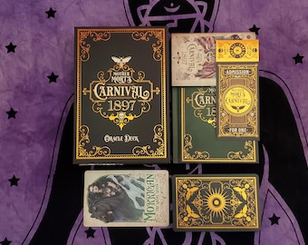 May include: An oracle deck with a black box featuring gold lettering and the text "Mother Mort's Carnival 1897". The image includes a green book, cards, and a ticket. The cards have illustrations and text, including "Morrigan the Great Queen".