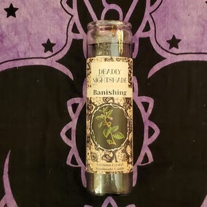 May include: A glass jar candle with a black wax fill. The label reads "DEADLY NIGHTSHADE Banishing" with a botanical illustration. The candle is set against a purple and black patterned background.