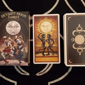 Può includere: Un mazzo di tarocchi Deviant Moon con una scatola e due mazzi di carte. La scatola presenta un'illustrazione surreale con una faccia di luna e figure in costume. Una carta mostra figure danzanti sotto un sole, etichettata "The Sun". L'altro mazzo ha un dorso decorativo.