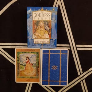 Könnte beinhalten: Ein Tarot-Deck mit dem Titel "The Goddess Tarot" von Kris Waldherr. Die Box ist blau mit einer Illustration einer Frau mit einer Eule. Zwei Karten werden angezeigt, eine mit einer Frau in einem gelben Kleid und die andere mit blauem Hintergrund und goldenen Linien.