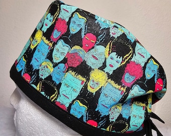May include: A black surgical scrub cap featuring a colorful pattern of classic monster faces in shades of blue, pink, yellow, and green. The cap has a black border and ties.