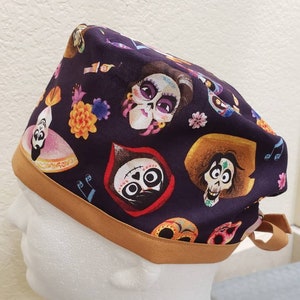 May include: A black scrub hat with a gold trim and a colorful print of characters from the movie Coco. The print features characters like Miguel, Hector, and Mama Imelda.
