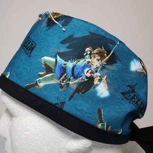 May include: A teal surgical scrub cap featuring a repeating pattern of a character from The Legend of Zelda: Breath of the Wild. The cap has a black band and ties. The text "The Legend of Zelda Breath of the Wild" is also visible.