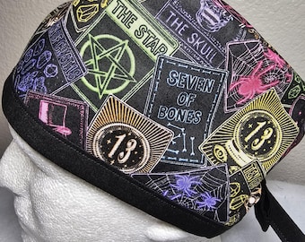 May include: A black scrub cap featuring a colorful tarot card design. The cap includes images of spiders, crystals, and the text "THE STAR", "THE SKULL", and "SEVEN OF BONES". The cap has a black brim and ties.