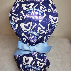 May include: Blue surgical scrub hat with a white bone pattern and the text "Ortho Surgery is Hip & Humerus".