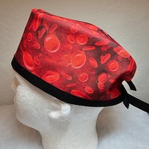 May include: Red and black surgical scrub hat with a red blood cell print. The hat has a black elastic band and ties at the back.