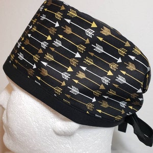 May include: Black scrub hat with a gold and white arrow pattern. The hat has a tie on the back.