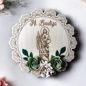 May include: A wooden round plaque with a lace border, featuring a saint image and the text "Mi Bautizo". The plaque is decorated with green and white paper flowers.