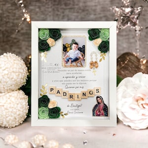 May include: A white framed photo display with a picture of a child, decorative paper flowers, and the word "PADRINOS" spelled out with letter tiles. The display includes text in Spanish and a small image of the Virgin Mary.