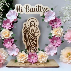 Puede incluir: Un letrero de madera con el texto "Mi Bautizo" en una fuente decorativa, rodeado de una corona de flores de papel rosa y blanco. Una imagen recortada de San Crist&oacute;bal se encuentra en el centro de la corona. El letrero tambi&eacute;n incluye el texto "Gianna Scarlet Cervantes Tamayo, Noviembre 23, 2024".