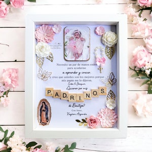 Puede incluir: Un marco de fotos blanco con fondo azul claro. El marco contiene una foto de una niña pequeña con un vestido rosa, una pequeña cruz blanca, un collar de cuentas blancas y un arreglo floral. El texto en el marco dice "Necesito un par de manos extra para ayudarme a aprender y crecer. y creo que ustedes son los mejores porque mis papás me lo dijeron. Erika & Joaquin ¿Quieren ser mis PAD₂RINOS de Bautigo? Con Amor, Kaylani Alejandre"