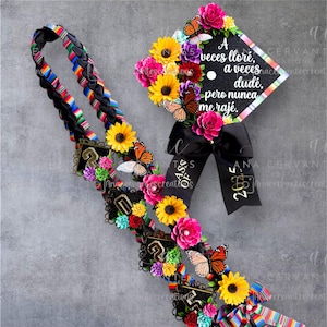 May include: A graduation lei and cap decorated with colorful flowers and butterflies. The lei features a braided design with a rainbow ribbon. The cap has the text "A veces lloré, a veces dudé, pero nunca me raje."