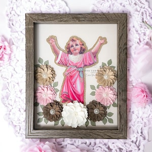 May include: A framed artwork featuring a vintage illustration of a young boy with outstretched arms, surrounded by a wreath of pink, brown, and white paper flowers. The artwork is titled "ANA CERVANTES CREATIONS".
