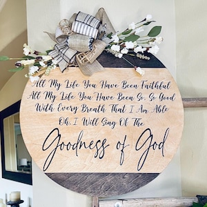 May include: A round wooden sign with the words "All My Life You Have Been Faithful...Goodness of God" in black script. It is decorated with a burlap and plaid bow, and white floral accents. The sign is a decorative piece.