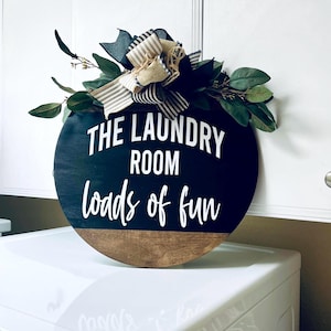May include: A round wooden sign with a black top and natural wood bottom. The text reads THE LAUNDRY ROOM loads of fun in white. A decorative bow made of black, white, and tan striped fabric sits on top with green faux leaves and branches.