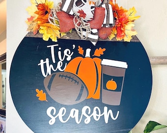 May include: A round wooden door sign with a dark blue background featuring the white text Tis' the season. The design includes a brown football, an orange pumpkin, and a brown coffee cup. A decorative bow with autumn leaves sits on top.
