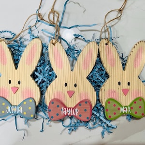 May include: Three wooden bunny ornaments with corrugated texture, pink painted cheeks, and black eyes. Each bunny features a bow tie with polka dots and a name written in white script: Joshua on a blue tie, Taylor on a red tie, and Max on a green tie.