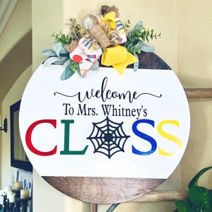 May include: A circular wooden sign with the words "welcome To Mrs. Whitney's CLASS" in various colours. The sign is decorated with a bow and greenery. The spiderweb in the word "CLASS" is black.