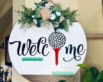 May include: A white round wooden sign with the words "Welcome" and a golf ball on a tee. The sign is decorated with a green, brown, and white bow.