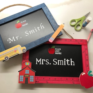 Puede incluir: Dos letreros de pizarra con marco de madera en rojo y azul. El letrero superior dice "Teach Inspire Mr. Smith" con una ilustración de autobús escolar amarillo. El letrero inferior dice "Teach Love Inspire Mrs. Smith" con una ilustración de una escuela roja.