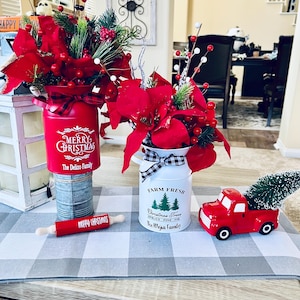 May include: Two Christmas themed centrepieces with red poinsettia flowers, greenery, and red berries. The centrepieces are in metal containers with the text "Merry Christmas" and "Farm Fresh Christmas Trees" with the family names below. A red toy truck with a small green Christmas tree in the bed is on a grey and white checked table runner.