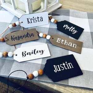 Puede incluir: Cinco etiquetas de regalo de madera con nombres escritos en ellas, "Krista", "Kasandra", "Ethan", "Molly" y "Justin". Las etiquetas están enhebradas con cuentas de madera y cuentas blancas.