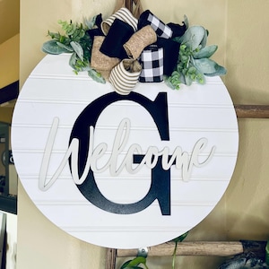 May include: A round white wooden door sign featuring horizontal shiplap lines and a large black letter G. The word Welcome is written in white script across the centre. A decorative bow made of burlap, black, and plaid ribbons sits on top.