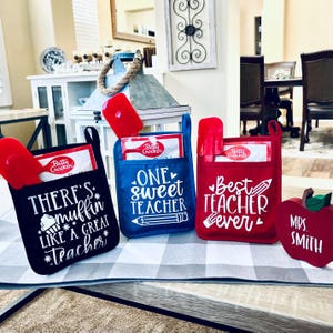 Op de afbeelding: Drie ovenwanten met verschillende spreuken erop. De eerste is zwart en zegt "There's muffin like a great teacher". De tweede is blauw en zegt "One sweet teacher". De derde is rood en zegt "Best teacher ever". Er is ook een rode appel met "Mrs. Smith" erop.