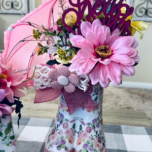 May include: Two pink rain boots with floral patterns are filled with pink and yellow flowers, a pink umbrella, and a wooden sign that says "Hello Spring".