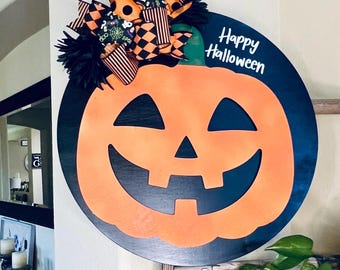 Puede incluir: Decoración redonda de Halloween con una cara de calabaza naranja sobre un fondo negro. La calabaza sonriente está adornada con un lazo de cintas negras, naranjas y blancas. Las palabras "Happy Halloween" están escritas en blanco.
