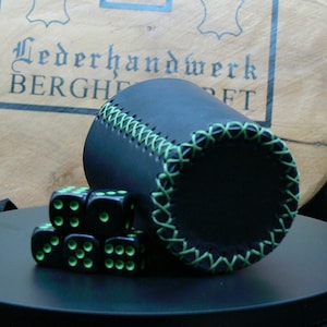 Könnte beinhalten: Ein schwarzer Leder-Würfelbecher mit grünen Nähten an den Rändern. Sechs schwarze Würfel mit grünen Punkten liegen vor dem Becher. Im Hintergrund ein Holzschild mit Text.